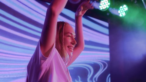 DJ plays music at night club party for crowd of cheerful people. Bright girl in spot light of multi-color neon. Smiling 20s dj mixing dance rhythm on mixer console. Energy dancer at concert stage show