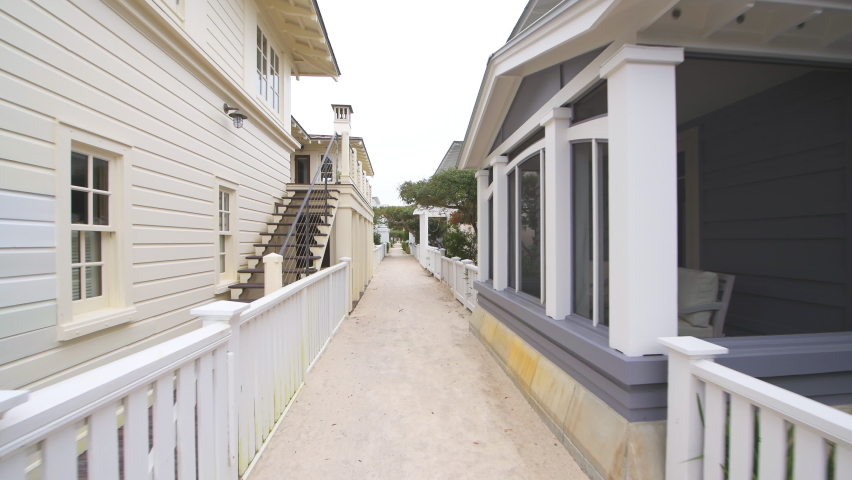 Pov point of view walking in Seaside, Florida by white beach wooden wood architecture house home real estate rental timeshare property with path alley by green landscaping shrubs, bushes sidewalk
