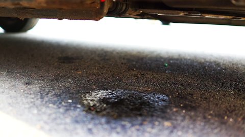 Parked car on street road with closeup of vehicle leaking fuel with puddle causing damage to asphalt due to rodents chewing cables