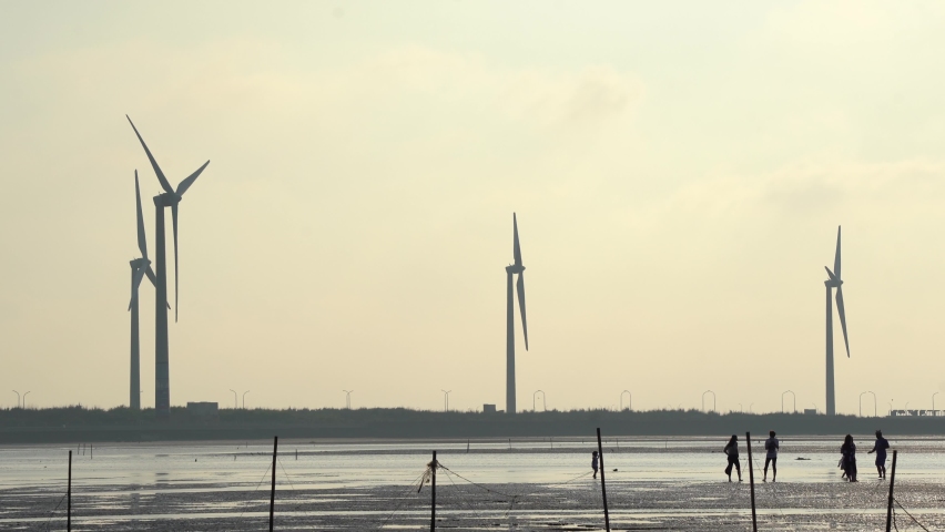 Silhouettes of tourists walking and playing on tidal flats with wind turbines spinning at the background, at Gaomei wetlands preservation area, Taichung, Taiwan.