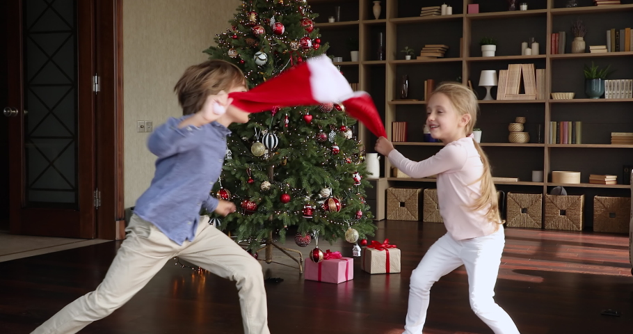 New Year is coming. Overjoyed little children jump laugh act funny fight using Santa caps at home. Excited preteen elder brother younger sister play have fun by decorated Xmas tree feel holiday spirit Royalty-Free Stock Footage #1080107843