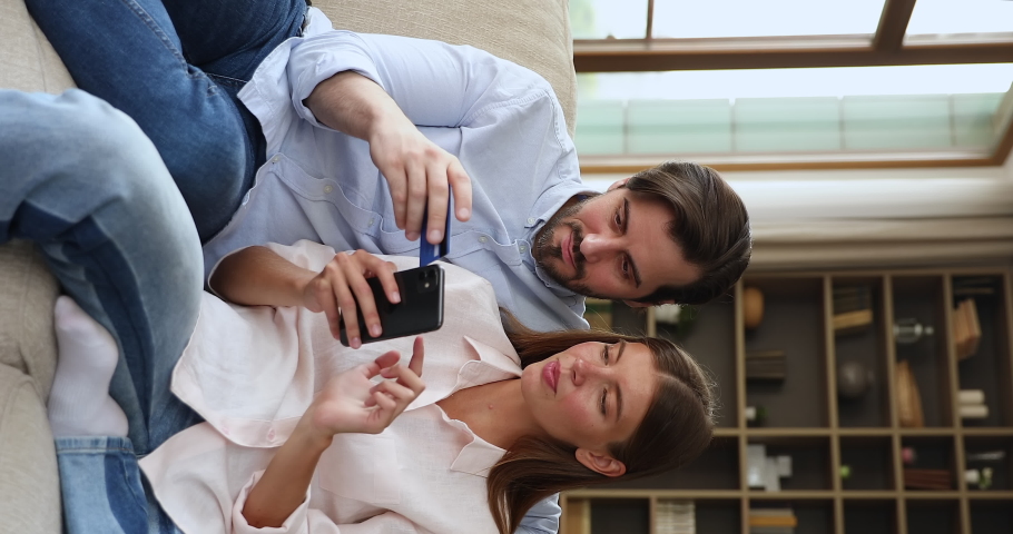 Vertical video happy millennial couple in love sit on sofa book tickets hotel order services make payment in mobile app using phone plastic card. Young spouses web bank customers check balance online