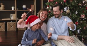 Celebrating New Year. Happy family mom dad preteen boy sit on warm floor under Christmas tree look at camera laugh light sparklers. Excited parents with son kid greet relatives with Xmas by video call