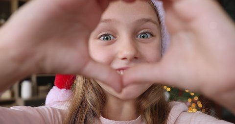 Excited little girl in Xmas cap video blogger make finger heart look at camera congratulate children audience with Christmas holidays. Happy small child shoot New Year video greeting card. Close up