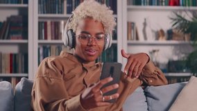 Happy young African American woman in wireless headphones holding mobile video call conversation with friends, talking chatting discussing life news distantly, resting on comfortable couch at home.