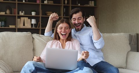 Excited young couple in love sit on couch read great news on laptop screen scream raise hands celebrate success. Happy emotional husband wife feel euphoric winning lottery prize getting loan approval