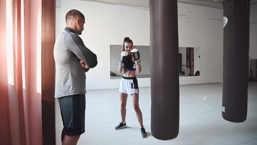 A strict trainer watches his female kickboxer student practice a punch on a punching bag in a spacious training room