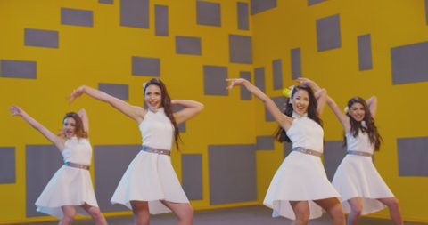 4K Group of beautiful  funny stylish girls in white same dresses dancing inside decorated pavilion with grey cubes and yellow wall background . Synchronized dance of female ensemble for video clip ,  