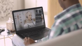 Telemedicine. Remote medicine concept. Back view of male patient having video call at home with female confident Arabic doctor on laptop, holding xray scan and consulting patient
