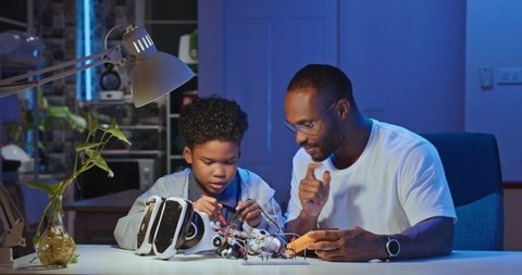 African american electronics engineer father and son sitting at table repairing a robot in living room home together at night. Future Science Education. People with technology or innovation concept.