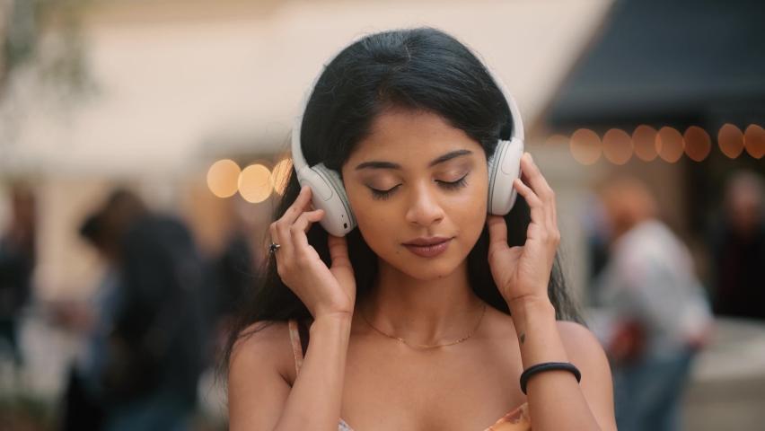 Closeup face of beautiful young Indian girl with long hair. Slow motion of smiling young girl listening to music in headphones at the city street.  | Shutterstock HD Video #1080124631