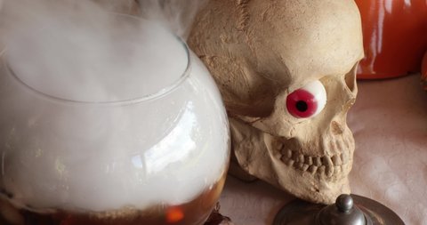 Smoke over glass bowl with Halloween decorations and realistic fake skull with red candy jelly eye. Preparing magic potion with fog made by dry ice put in dark liquid. Halloween potion making concept