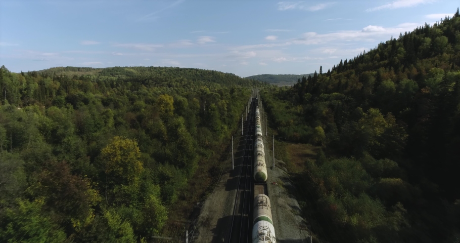Freight long train carries with oil tank and petrol carriages an electric locomotive by Trans Siberian railways under the rock and near mountain river. Aerial drone wide view at summer sunny day Royalty-Free Stock Footage #1080127298