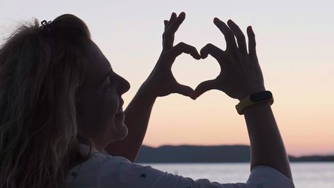 Silhouette of a woman with long hair holding her hands and fingers joined in a heart shape, on the sea shore, in the evening. Handheld, real time, Summer, vacation, happiness, love, romance.