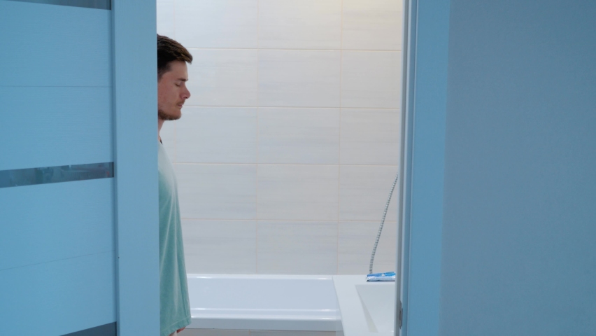 The man in the bathroom goes to the sink and starts brushing his teeth. Side view. Overall plan