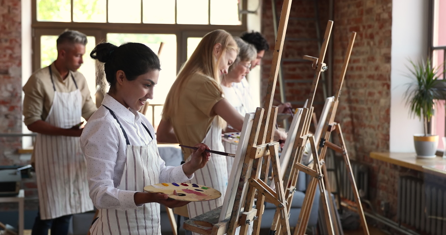 Young inspired Indian student woman attend in painting class, stand in front of easel holds paintbrush and palette makes strokes, absorbed in creative process. Artistic education, arts studio concept Royalty-Free Stock Footage #1080128594