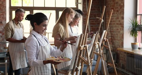 Young inspired Indian student woman attend in painting class, stand in front of easel holds paintbrush and palette makes strokes, absorbed in creative process. Artistic education, arts studio concept