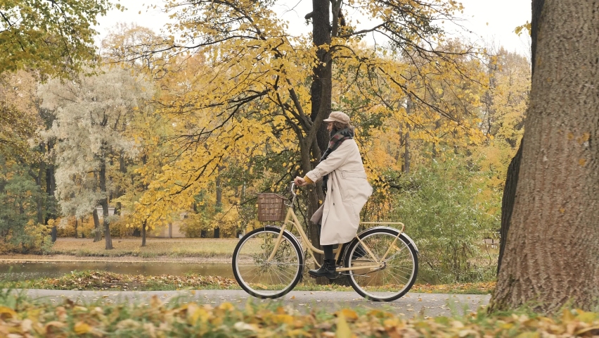 Young stylish woman in a beige raincoat rides a city bike with a basket through an autumn park in the middle of yellow trees and leaf fall, in slow motion, side view