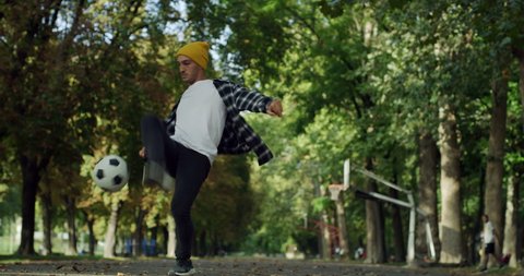 Cinematic shot of young carefree arab freestyle soccer player is juggling sport ball with his knee and feet while training tricks of self-expression with football in public green park.