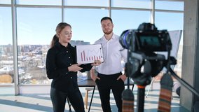 Attractive girl next to a young businessman records video. Young woman holding paper with graphs and speaking in front of professional camera in office room.