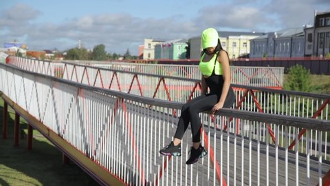 A young woman with dark hair in tight-fitting sportswear sits on a bridge.