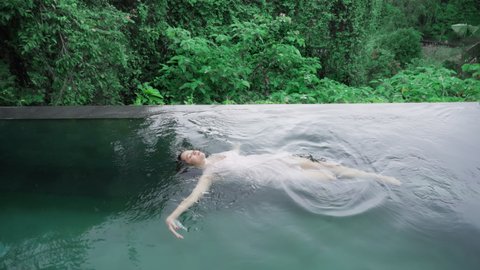 Top shot view of a beautiful girl in a soft pink evening dress who is swimming on her back in a pool with emerald green water in cloudy weather. Woman is lying on her back in a pool. 