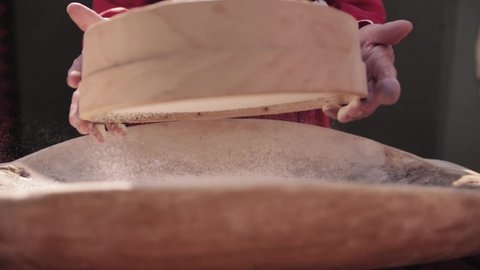 Elderly woman is sifting flour through wooden sieve for baking. White powder flying in different directions. Preparing flour to baking. Close up. Slow motion.