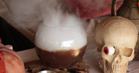 Smoke over glass bowl with Halloween decorations and realistic fake skull with red candy jelly eye. Witch hands prepare magic potion and put spell. Halloween witchcraft and potion making concept