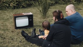 Family of a man, a woman and little baby are sitting on the grass and watching an old retro TV. They clap their hands. TV is strobing. The concept of post apocalypse, horror. Dependence on television