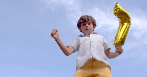 A cute 7-8 years old boy having fun jumping at trampoline with an inflatable number seven on the blue sky background, 4k slow motion low angle view