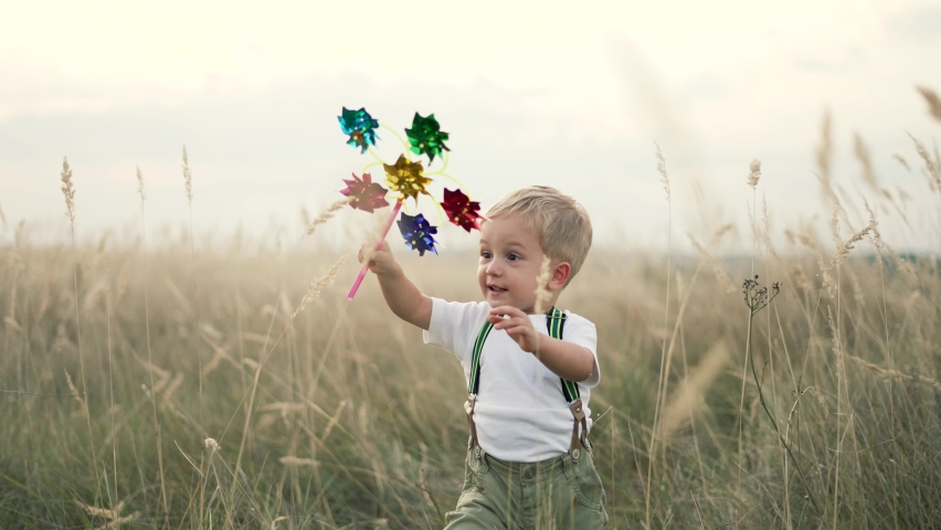 Happy kid with toy in park. Toy windmill in hand of child. Happy kid run over spikelets. Little hand of child is holding toy windmill. Kid plays in park. Happy childhood concept. Happy kid run in park Royalty-Free Stock Footage #1080170486