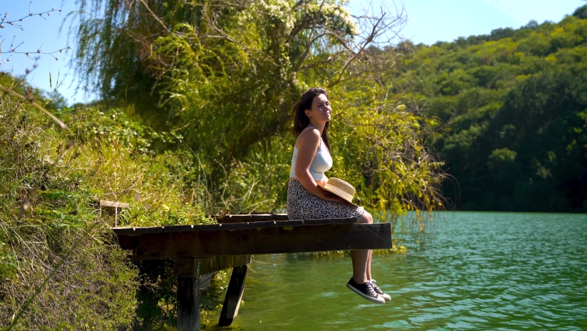 4k video. A young woman is sitting on a wooden pier on the emerald lake in the forest and cheerfully wiggles her legs admiring the view
