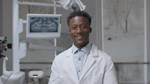 Professional african american male doctor dentist taking off protective eyehlasses and smiling widely to camera, sitting at modern office, slow motion