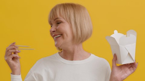 Happy Senior Woman Holding Paper Lunch Box And Chopsticks, Smiling Mature Female Recommending Chinese Asian Food Delivery While Standing Over Yellow Studio Background, Slow Motion Footage