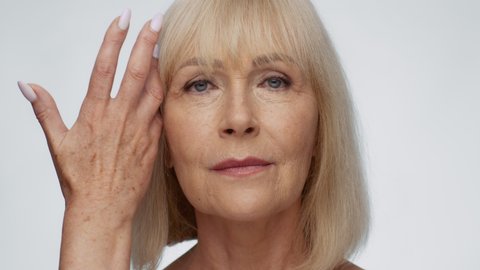 Female beauty. Close up portrait of confident attractive senior woman with wrinkled skin looking at camera, caressing her face with hand with dark spots, white studio background, slow motion