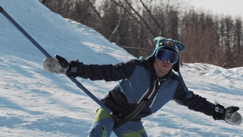 Happy snowboarder climbs a hill on a T-bar lift and performs a frontside 180 buttering trick on a sunny day at a ski resort. Climb to the top of the slope on a rope lift.