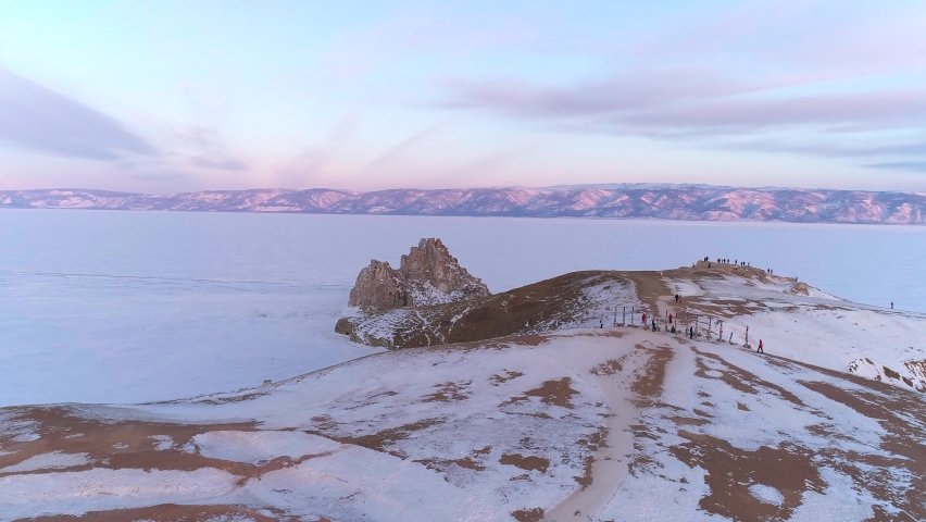 Aerial over cape Burkhan and Shamanka rock on Olkhon island at beautiful pink sunrise. Tourists walking around the ritual wooden pillars. Famous tourist spot on Baikal. Winter landscape of lake Baikal