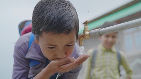 close up shot of a young little school boy with backpack drinking clean water from a running tap in a rural village. Concept of a natural resources conservation