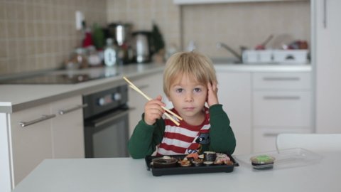 Cute blond child Eat Sushi Rolls At Home. Happy boy ready for eating sushi, sitting in a sunny kitchen