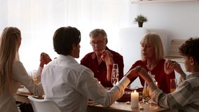 Family Celebrating Thanksgiving Day Praying Together Holding Hands With Eyes Closed Before Celebratory Dinner Sitting At Table In Modern Kitchen At Home. Autumn Holidays Celebration Concept