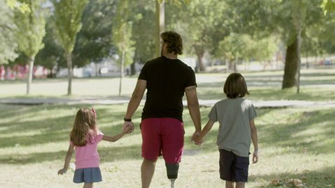 Back view of dad with artificial leg walking with kids in park. Father going to amusement park with daughter and son during summer holiday, spending time together. Disability, family concept