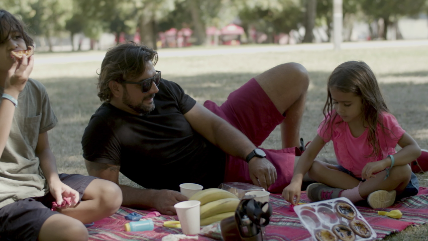 Single father having picnic with kids in nature. Cute girl blowing soap bubbles, funny boy enjoying taste of cake, dad with prosthesis leg lying on blanket. Spending time together, disability concept