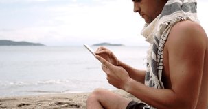 Caucasian male sitting on beach scrolling on digital tablet 