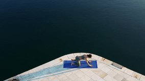 Video of caucasian female in active wear doing exercises near calm water surface recreating and keeping perfect body shape, aerial view of woman having morning yoga near ocean