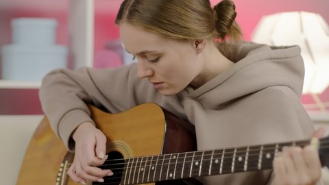 Excited girl playing acoustic guitar in living room. Happy musician playing chords on string instrument. Musician performing musical composition on guitar.