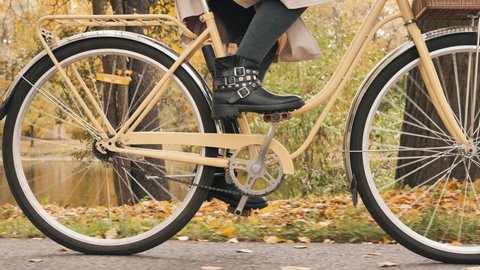 Women's legs in fashionable boots are pedaling a city bike in the autumn park in slow motion, cycling through the autumn city park