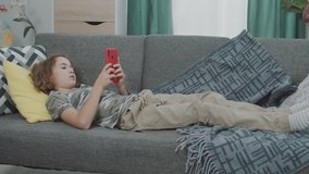 Handsome and Cute Young Caucasian Boy Laying on a Grey Sofa, Playing Games on his Red Smartphone and Falling Asleep Slowly, Head on a Yellow Pillow. Wearing Cozy Brown Pants and a Camouflage Shirt.