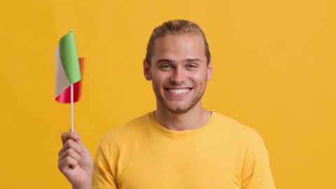 Joyful Young Handsome Guy Waving With Flag Of Italy And Looking At Camera, Cheerful Millennial Blonde Guy Holding Italian National Ensign While Posing Over Yellow Background, Slow Motion Footage