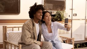 Two Multiethnic Friends Making Video Call With Smartphone At Campsite, Young Asian Woman And Black Guy Sitting Near Modern Camper Van, Waving Hands At Camera And Sending Air Kisses, Slow Motion