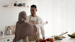 Happy Islamic Couple Hugging Expressing Love Cooking Together Standing In Modern Kitchen At Home. Husband Embracing Wife In Hijab. Modern Muslim Family Concept. Slowmo